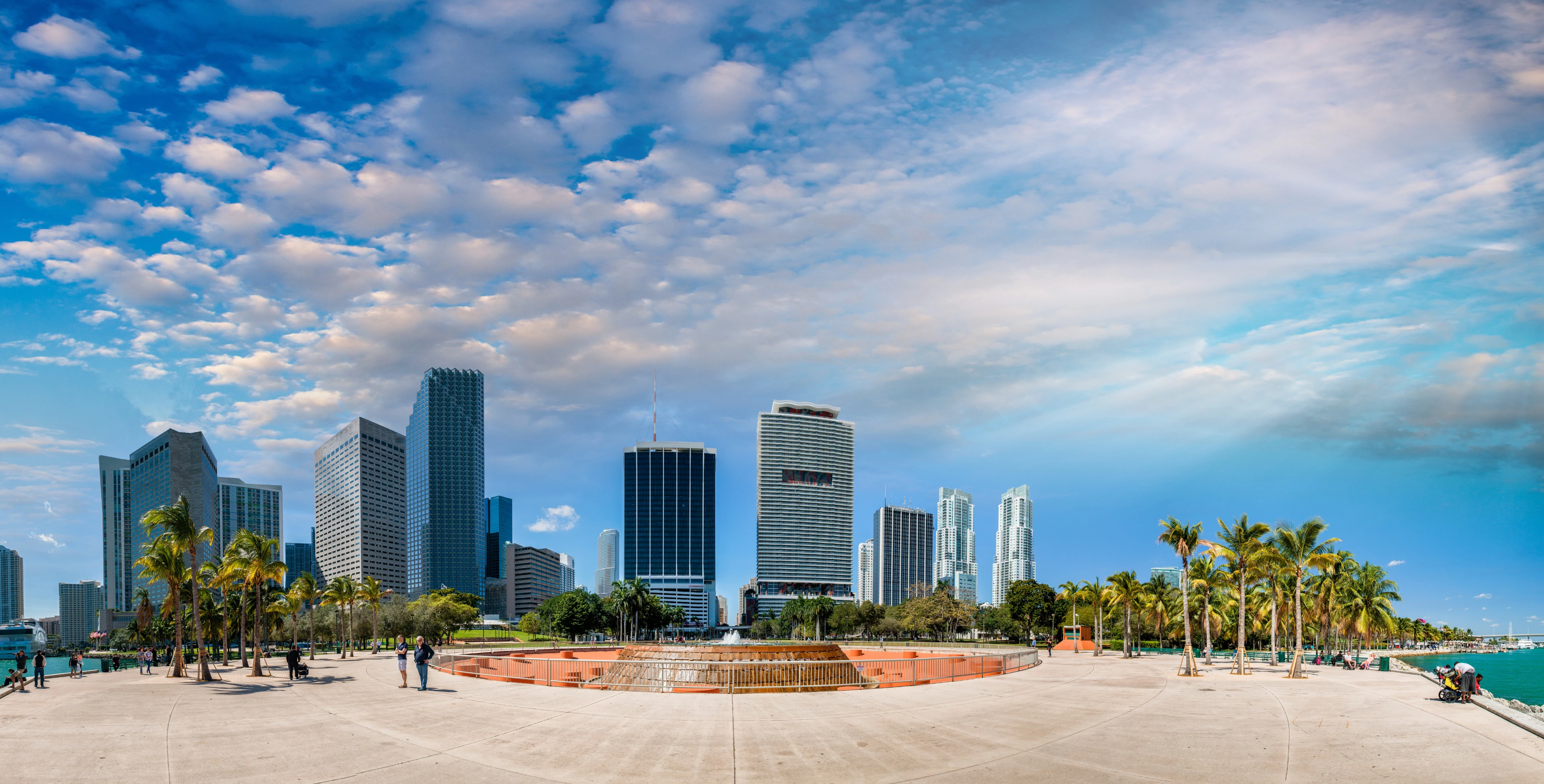 Imagem do Bayfront Park em Miami, mostrando um espaço amplo com palmeiras e uma fonte ao centro, cercado por arranha-céus que compõem o skyline da cidade. A cena transmite uma sensação de tranquilidade e integração entre natureza e cidade, em um dos parques mais emblemáticos de Miami.