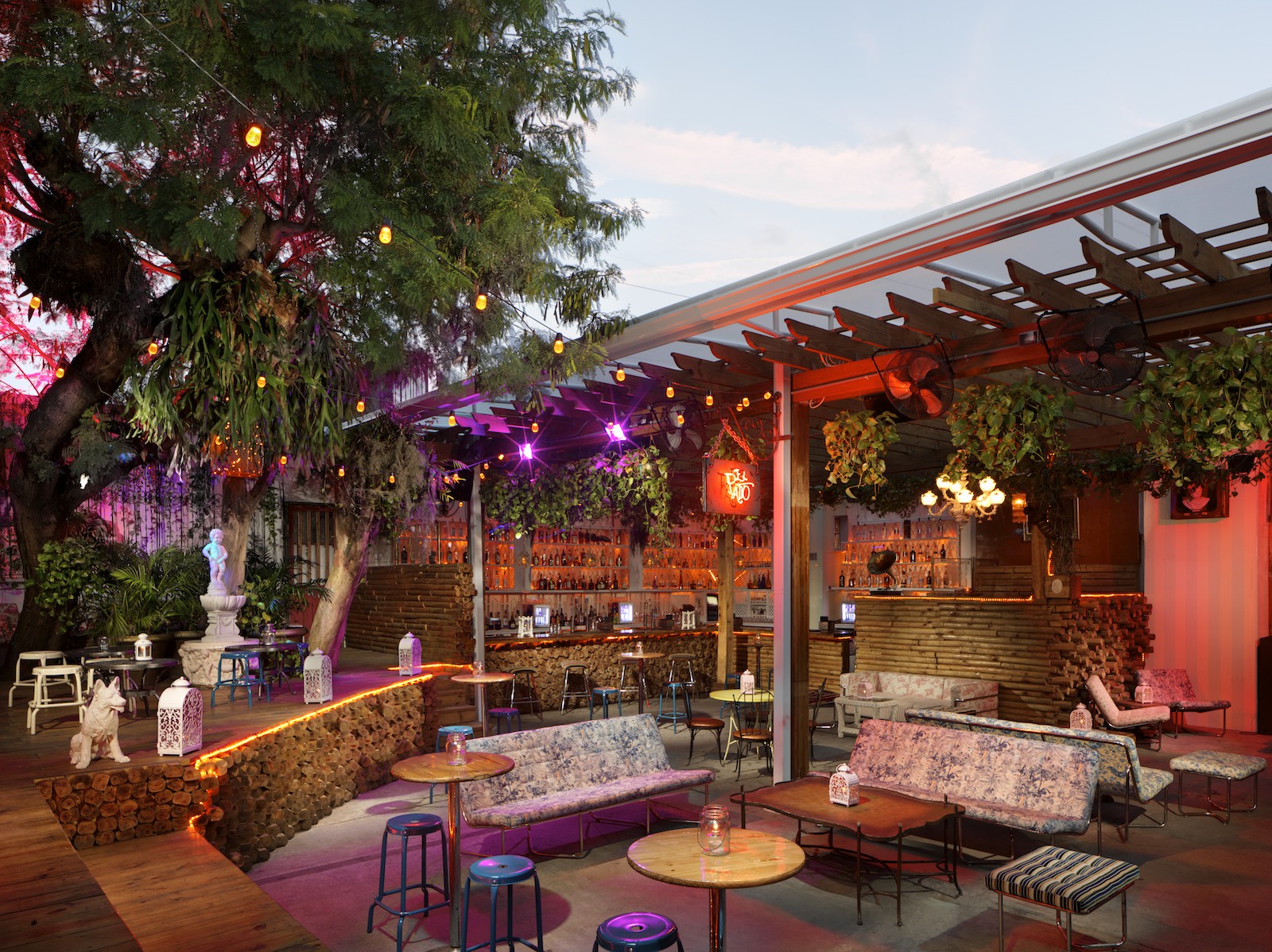 Área externa da El Patio em Miami, mostrando um ambiente com decoração charmosa e rústica. Móveis acolhedores, plantas pendentes e luzes decorativas criam uma atmosfera intimista e convidativa. O bar ao fundo está iluminado, completando o cenário perfeito para uma noite descontraída.