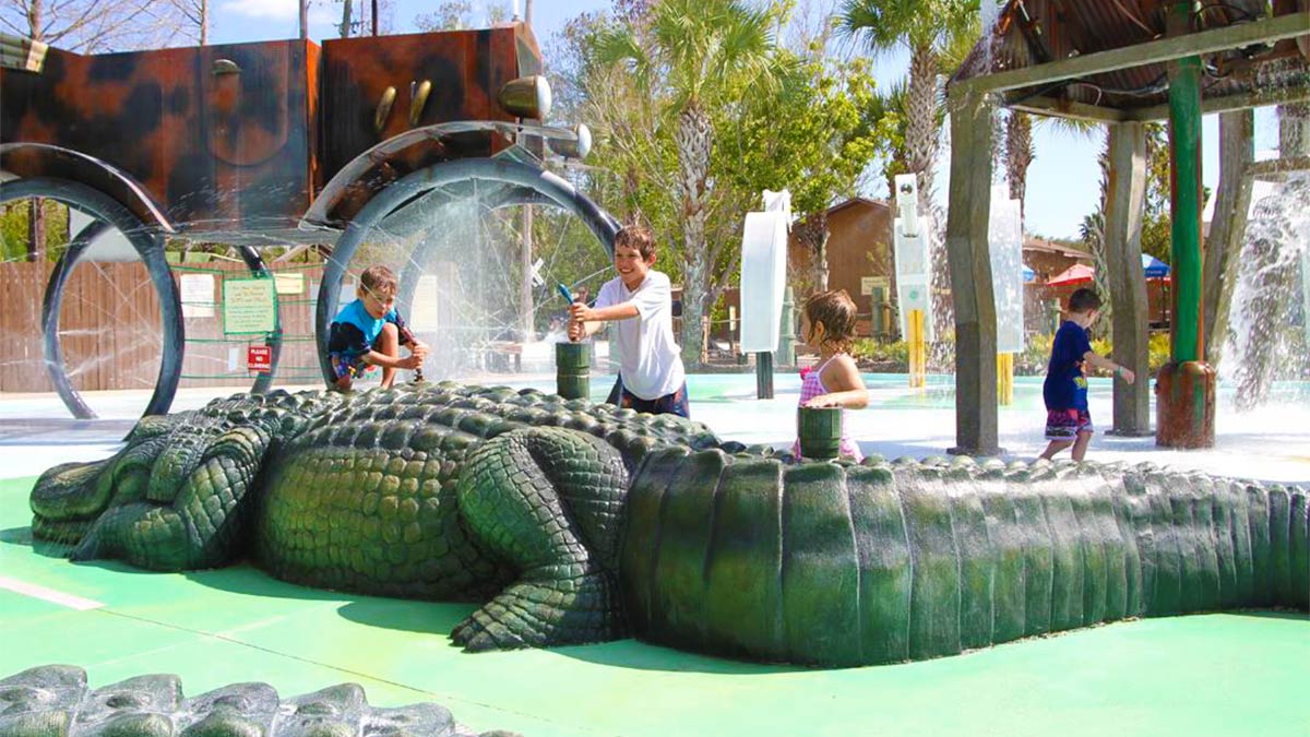 Crianças brincando com uma escultura de jacaré em Gatorland, Orlando, em um ambiente de parque aquático ao ar livre.