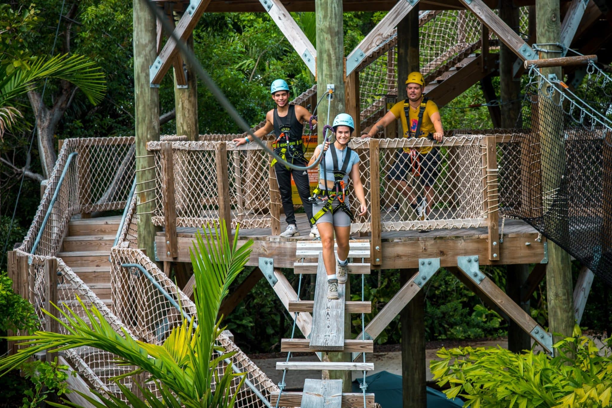 Pessoas se divertindo na Jungle Island em Miami, participando de uma atividade de tirolesa em uma plataforma de madeira cercada por redes de segurança. Eles estão usando equipamentos de proteção e capacetes, prontos para explorar a atração em meio à vegetação tropical. A cena transmite uma atmosfera de aventura e diversão ao ar livre.