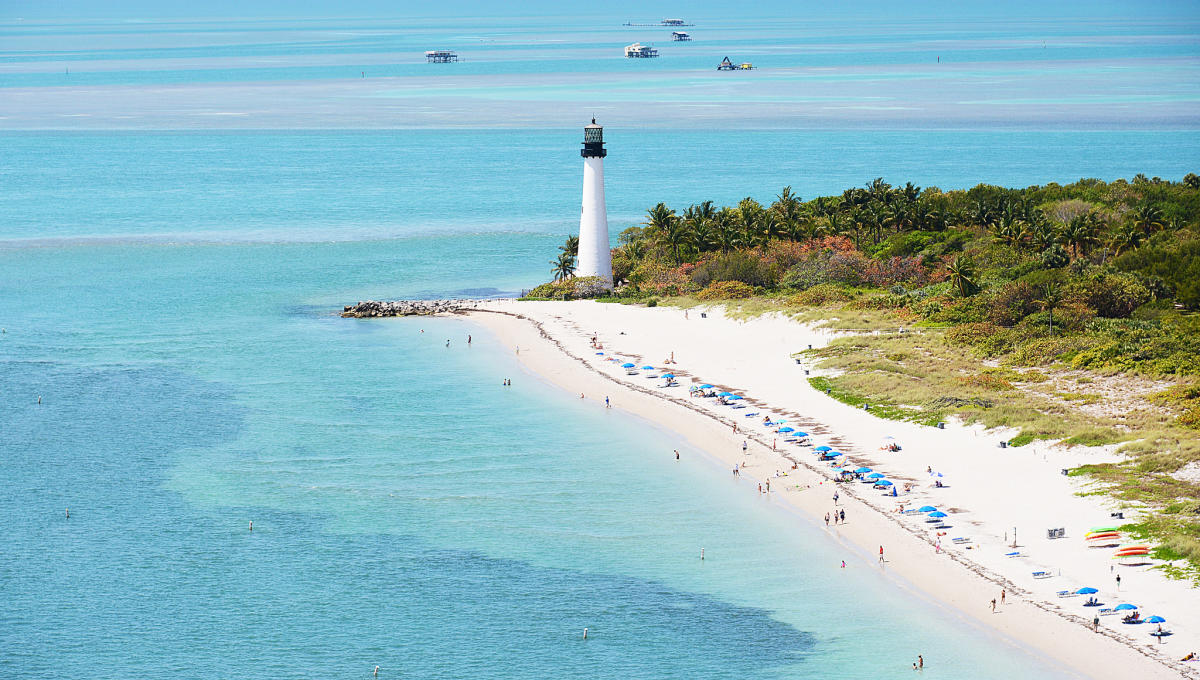 Imagem aérea de Key Biscayne em Miami, mostrando uma praia de areia branca com guarda-sóis azuis ao longo da costa. O farol histórico está situado ao fundo, rodeado por vegetação tropical e águas azuis e claras, proporcionando um cenário tranquilo e pitoresco para os visitantes desfrutarem do local.