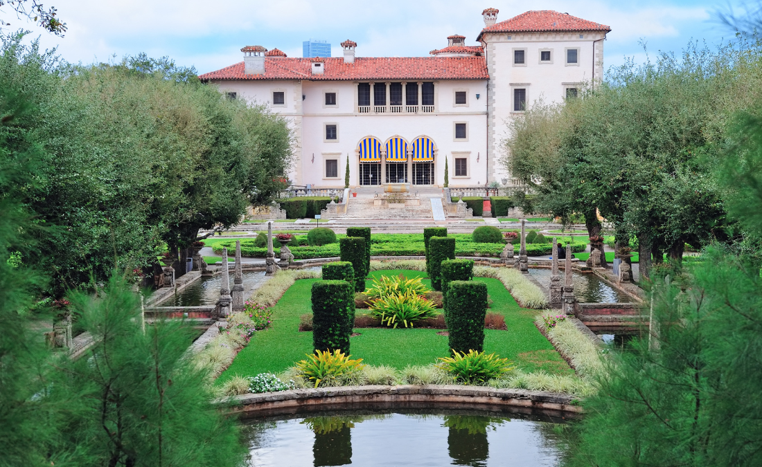 Vista frontal do Vizcaya Museum and Gardens em Miami, mostrando os jardins bem cuidados, decorados com fontes, arbustos geométricos e canteiros de flores. O palácio ao fundo, de estilo renascentista, cria um ambiente elegante e encantador, ideal para passeios em família e momentos de relaxamento em meio à beleza natural e histórica.