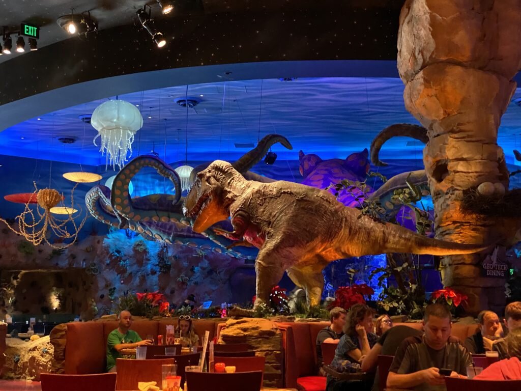 Interior do T-REX Café em Orlando, mostrando um dinossauro Tiranossauro Rex em destaque, cercado por elementos de decoração subaquáticos e mesas ocupadas por clientes.