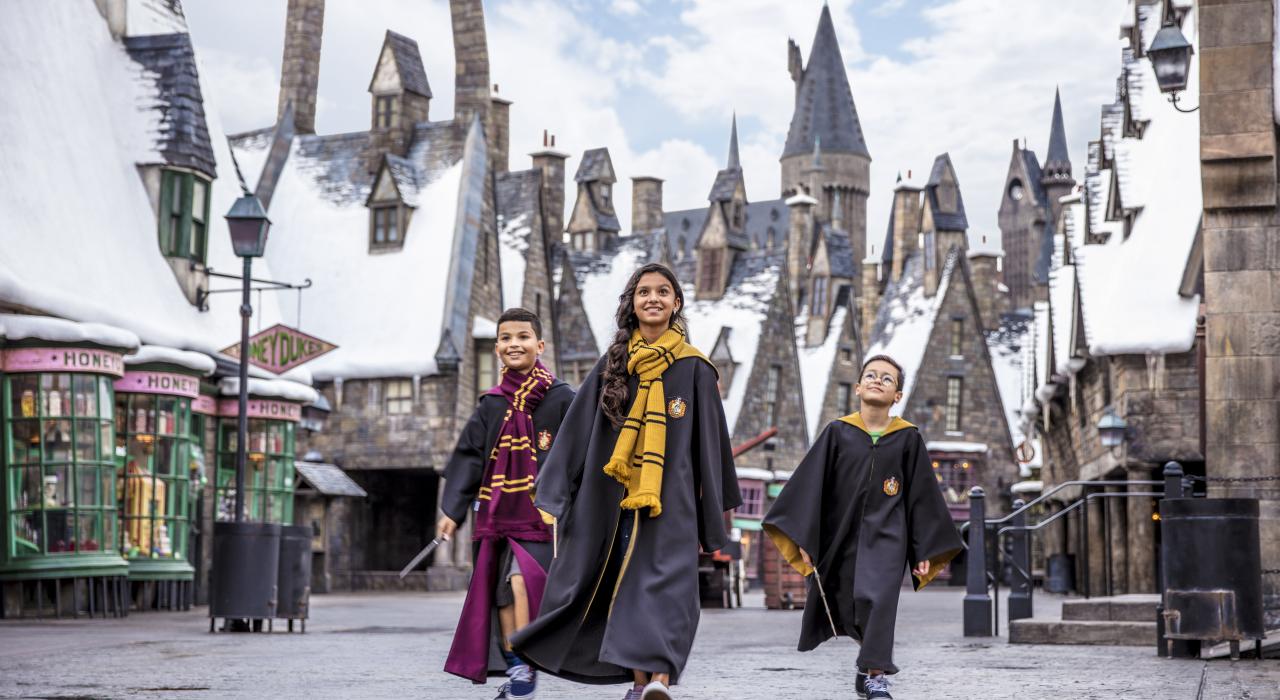 Três crianças vestindo trajes de Hogwarts caminham pelo The Wizarding World of Harry Potter no Universal Orlando Resort, cercadas por prédios temáticos cobertos de neve.