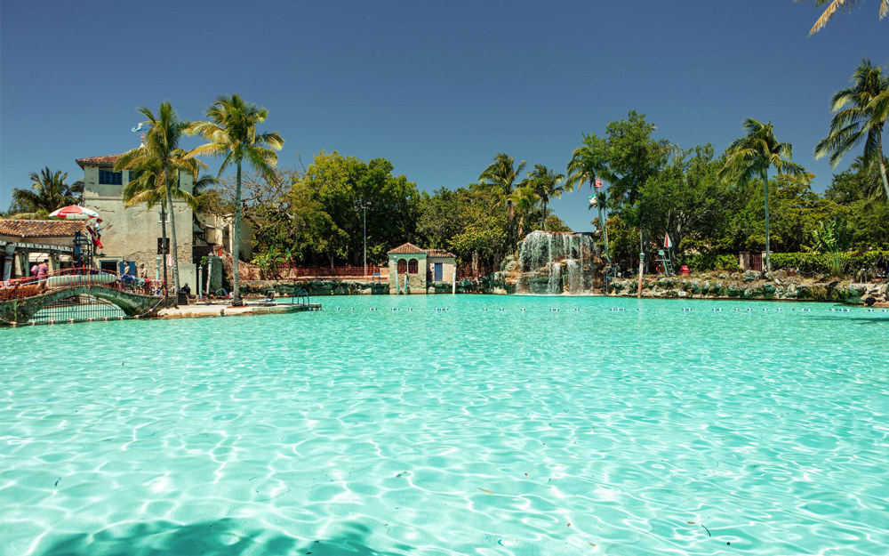 Imagem da Venetian Pool em Miami, mostrando uma piscina ampla de águas azul-turquesa cercada por palmeiras, com uma cachoeira ao fundo. A piscina, que lembra um oásis, é rodeada por uma arquitetura em estilo mediterrâneo, criando uma atmosfera charmosa e relaxante, ideal para um dia em família ou entre amigos.