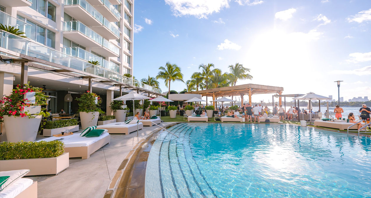 Área externa da Baia Beach Club em Miami, mostrando uma piscina rodeada por espreguiçadeiras confortáveis e guarda-sóis brancos, com pessoas relaxando sob o sol. Ao fundo, há uma vista da baía e palmeiras que complementam a atmosfera tropical e luxuosa do local.