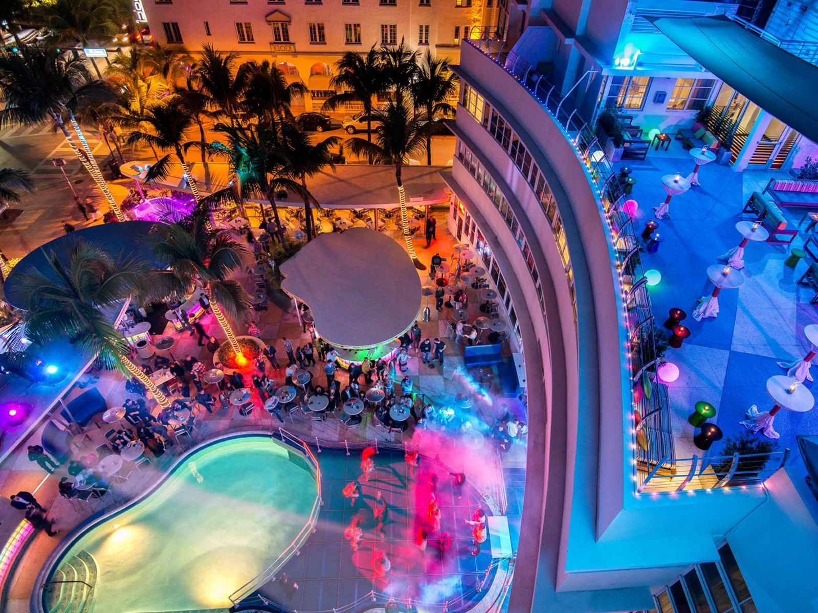 Vista aérea da The Clevelander em Miami à noite, destacando a piscina em formato orgânico, rodeada por mesas, palmeiras e luzes coloridas que iluminam o ambiente. A multidão está reunida ao redor, aproveitando a atmosfera festiva e as diversas áreas de entretenimento, em um local cheio de movimento e energia.