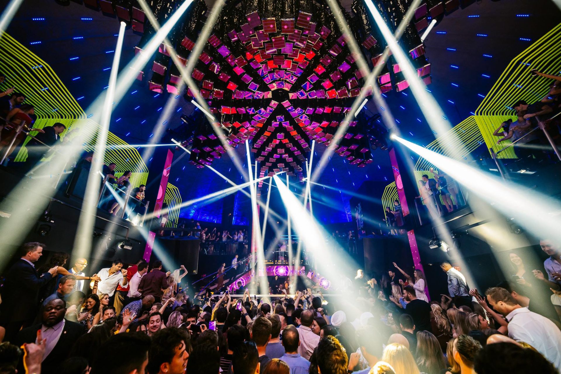 Boate LIV Nightclub em Miami lotada, com uma multidão dançando e luzes de palco intensas iluminando o ambiente. O teto apresenta uma estrutura geométrica iluminada, complementando a experiência visual e criando uma atmosfera de festa vibrante.