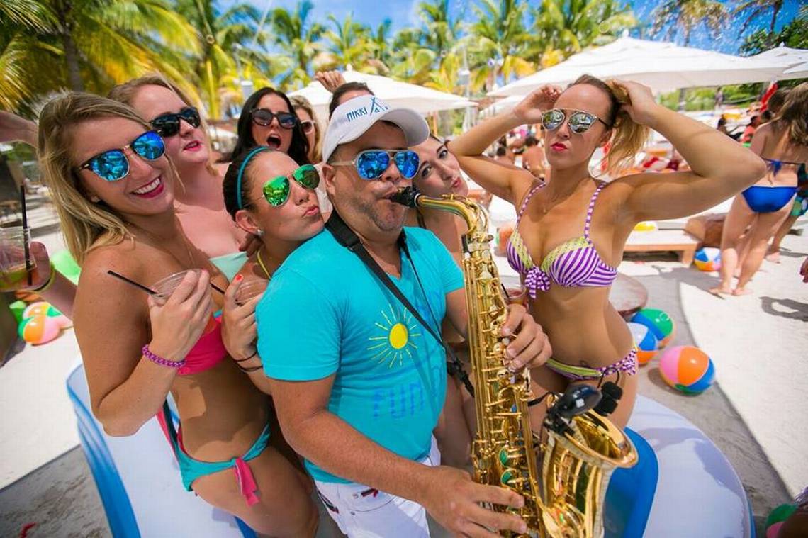 Músico tocando saxofone durante uma festa ao ar livre na Nikki Beach em Miami, cercado por mulheres em trajes de banho que sorriem e posam para fotos. O ambiente está decorado com palmeiras e espreguiçadeiras, criando uma atmosfera de diversão tropical e relaxamento na praia.