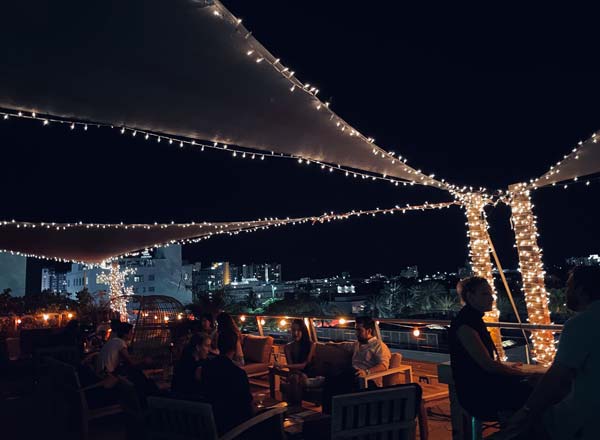 Área ao ar livre da Sky Yard Nightclub em Miami, decorada com luzes de festa que criam uma atmosfera intimista e charmosa. Grupos de pessoas estão sentados em mesas, aproveitando a noite, com a vista da cidade ao fundo.