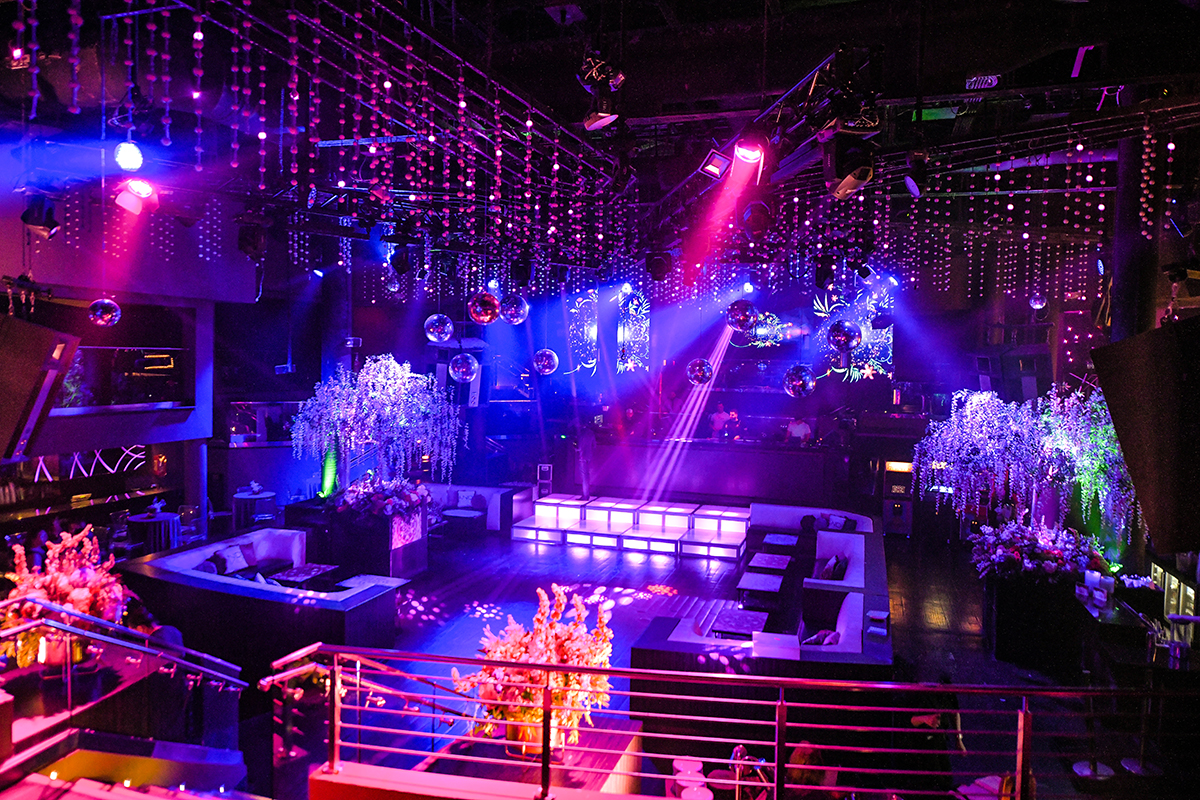 Interior da boate Story Nightclub em Miami, mostrando uma decoração elegante com luzes em tons de rosa, azul e roxo, espelhos brilhantes pendurados no teto, e arranjos de flores decorativos ao redor da pista de dança e áreas de descanso. O ambiente está pronto para receber o público e proporcionar uma noite luxuosa e animada.