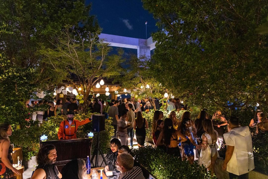 Sugar Rooftop em Miami durante a noite, mostrando uma multidão de pessoas se divertindo em meio a vegetação exuberante e luzes pendentes que criam uma atmosfera acolhedora e festiva. As pessoas estão interagindo em grupos, conversando e aproveitando a música, em um espaço ao ar livre decorado com estilo natural.