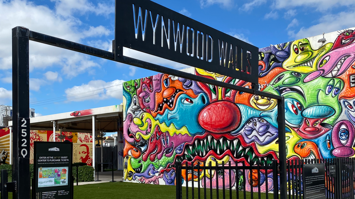 Imagem da entrada do Wynwood Walls em Miami, com um mural de arte urbana colorido ao fundo. A placa 'Wynwood Walls' está em destaque, enquanto o mural mostra uma mistura de formas e cores vivas, representando a expressão artística característica do bairro. Wynwood é conhecido por suas paredes grafitadas que atraem turistas e amantes da arte.